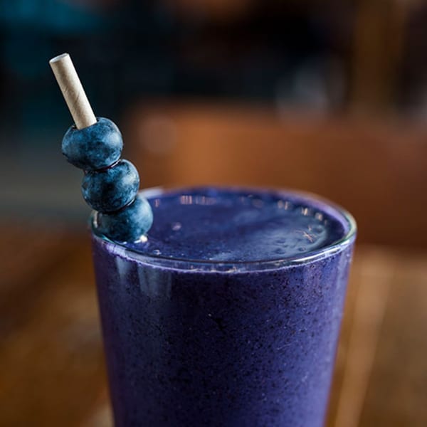 A vibrant blueberry smoothie in a glass, garnished with three fresh blueberries on a paper straw.