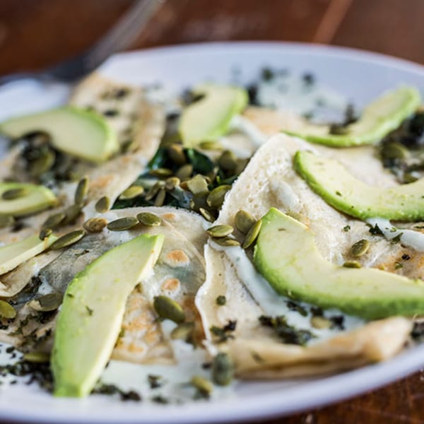 Vegetarian crepes topped with sliced avocado, pumpkin seeds, and a creamy sauce on a white plate.