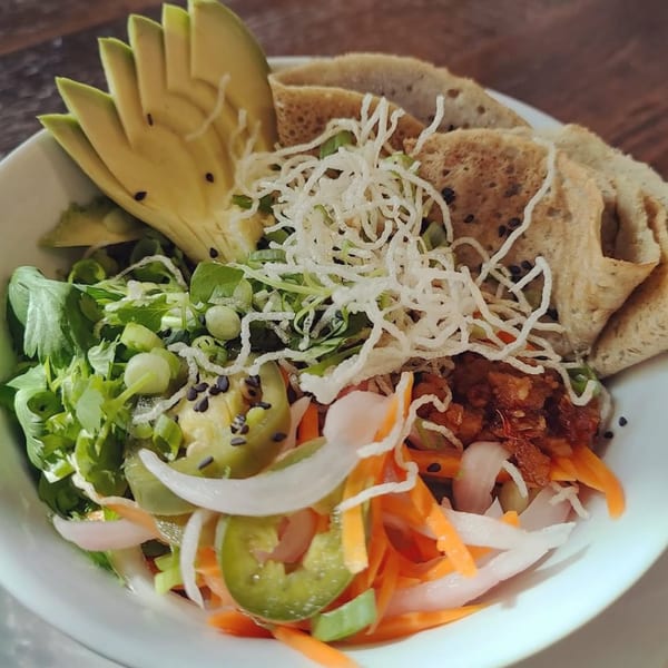 A vibrant vegetarian bowl with sliced avocado, crispy noodles, greens, and pickled vegetables.