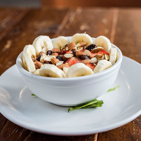 A white bowl of sliced bananas, strawberries, almonds, and raisins on a white plate.