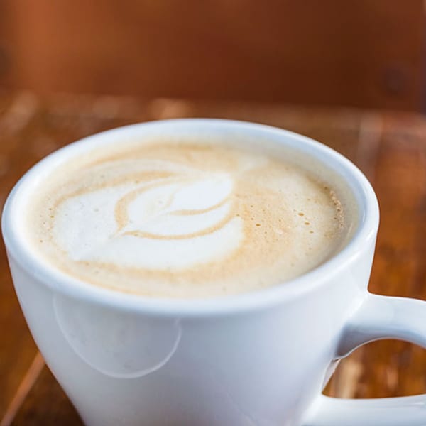 A white mug of latte with a leaf design in the foam on a wooden table.